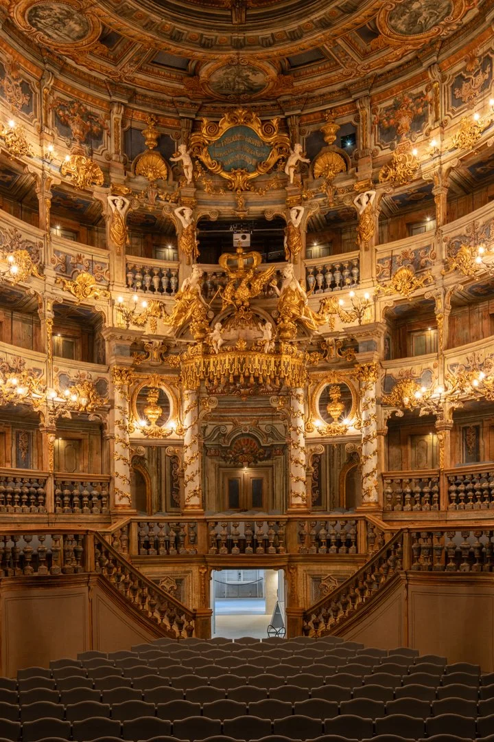 Interior of an ornate opera house or theater with a richly decorated stage, featuring gold accents, intricate woodwork, sculptures, chandeliers, and multiple tiers of balconies.