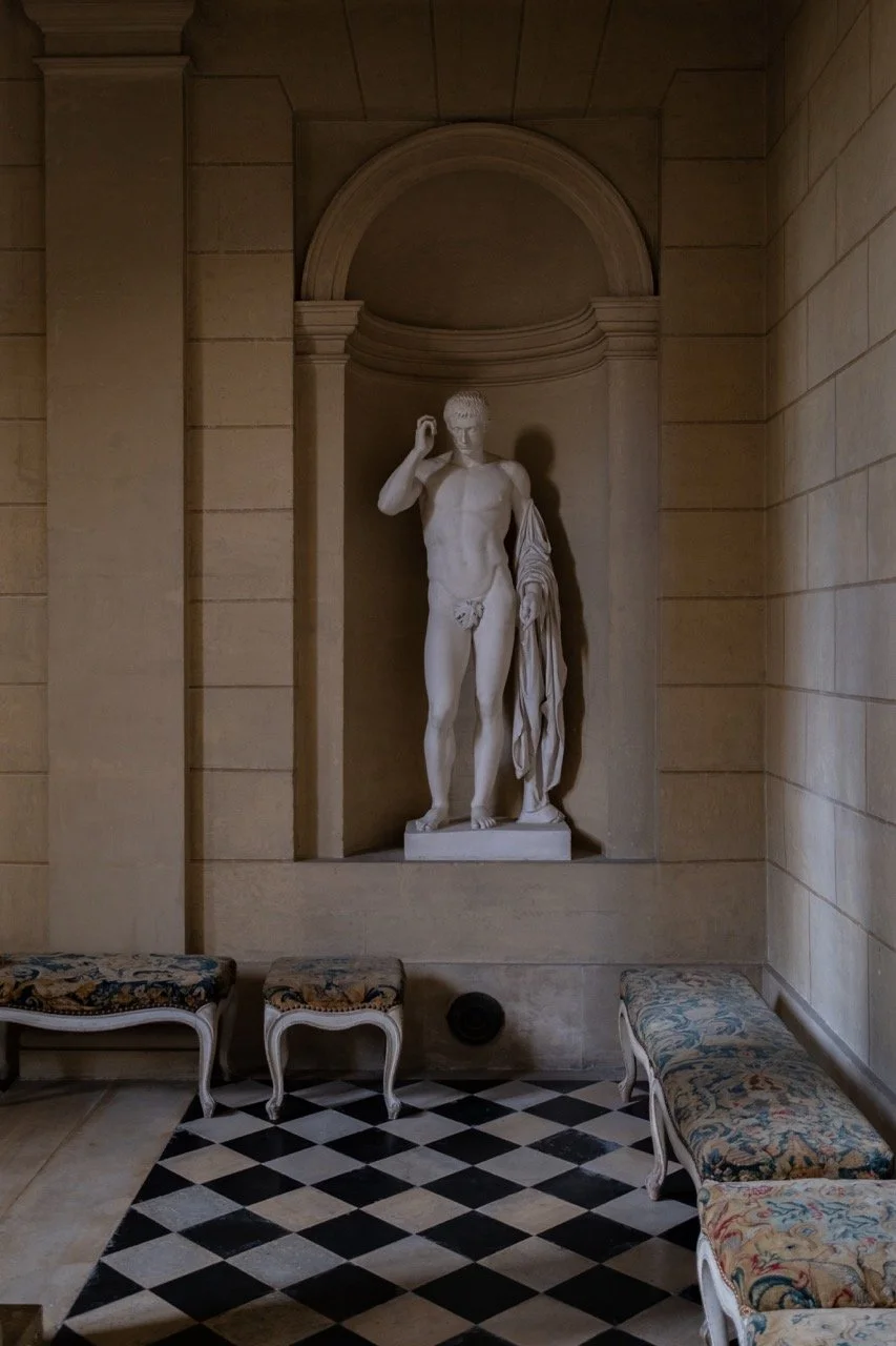 Classical marble statue of a nude male figure with a cloth draped over one arm, standing in a display niche with an arched top, in a room with patterned benches and a black and white checkered floor.