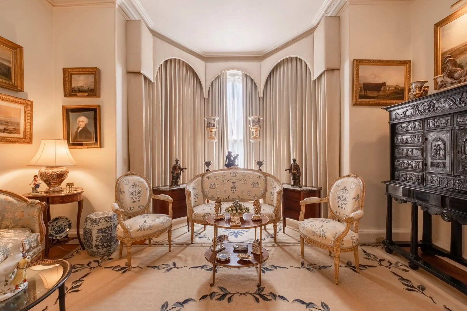 Decorative living room with antique furniture, ornate curtains, framed artwork, a patterned rug, and various decorative items including sculptures and a lamp.