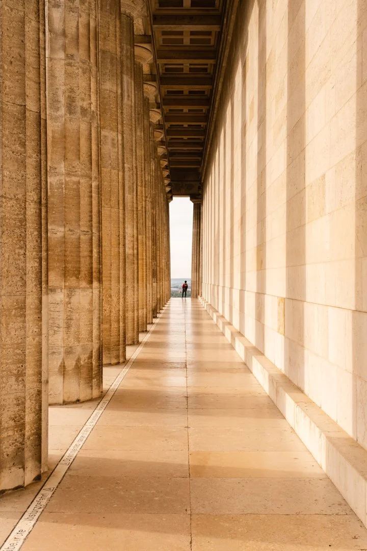 A person standing at the end of a colonnaded walkway between ancient Greek-style stone columns and a stone wall, with an open view of the sky and distant landscape.