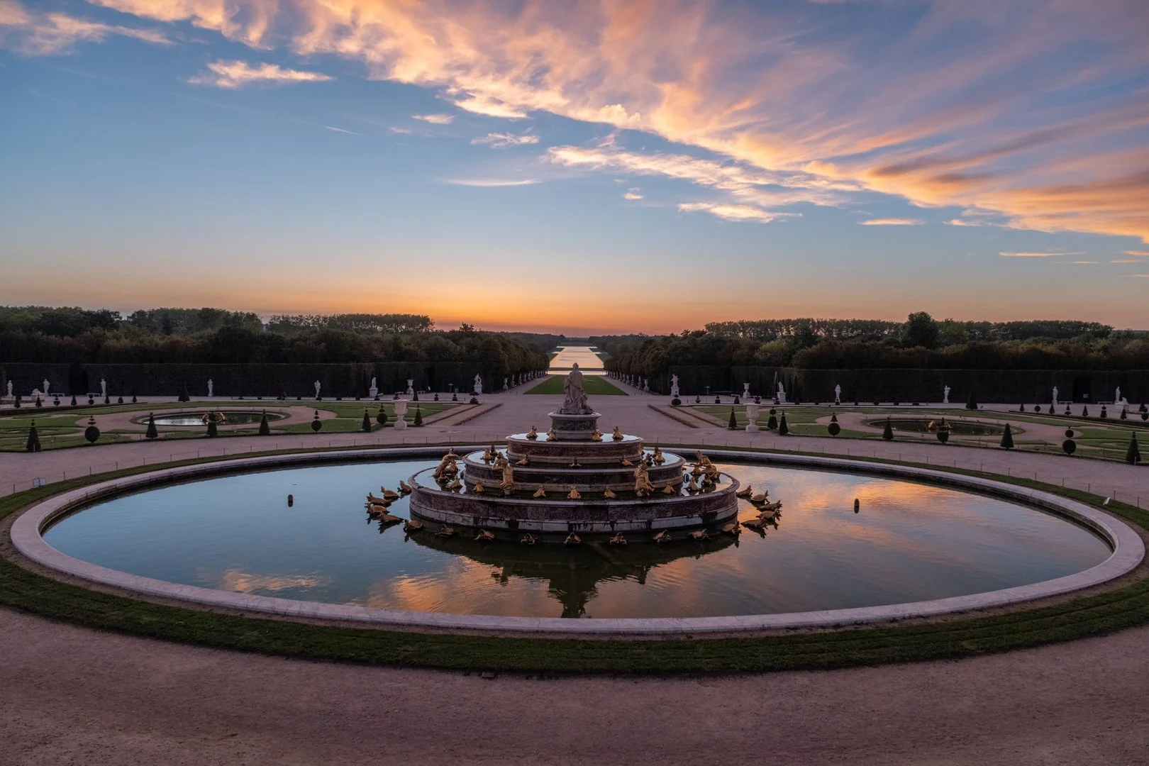 A large ornamental fountain with statues surrounds the top with a serene reflecting pool, set in a symmetrical garden at sunset, with a sky filled with wispy clouds.