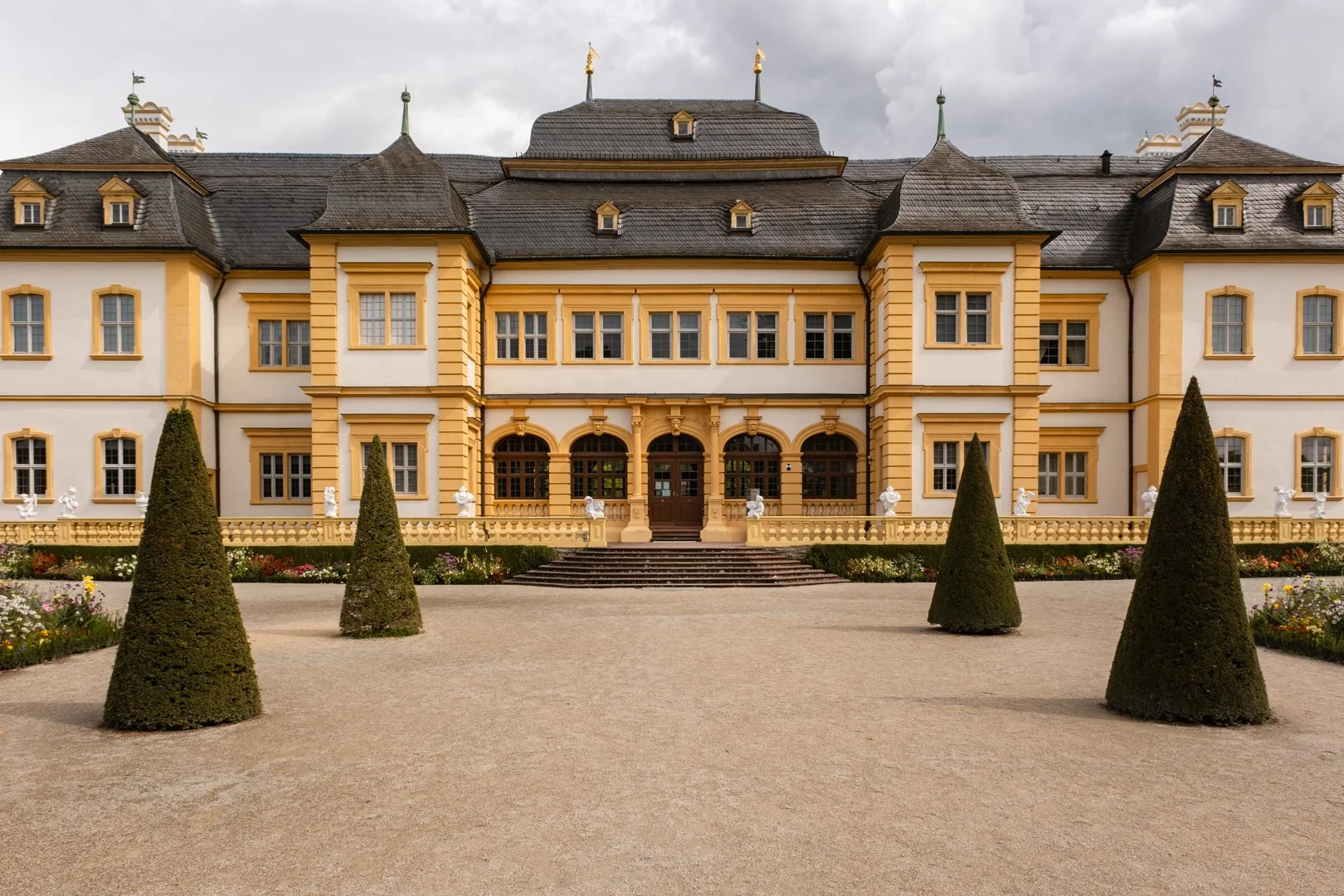 A large yellow and white mansion with ornate architecture, surrounded by a landscaped garden with cone-shaped bushes and statues, and stairs leading to a central entrance under an overcast sky.