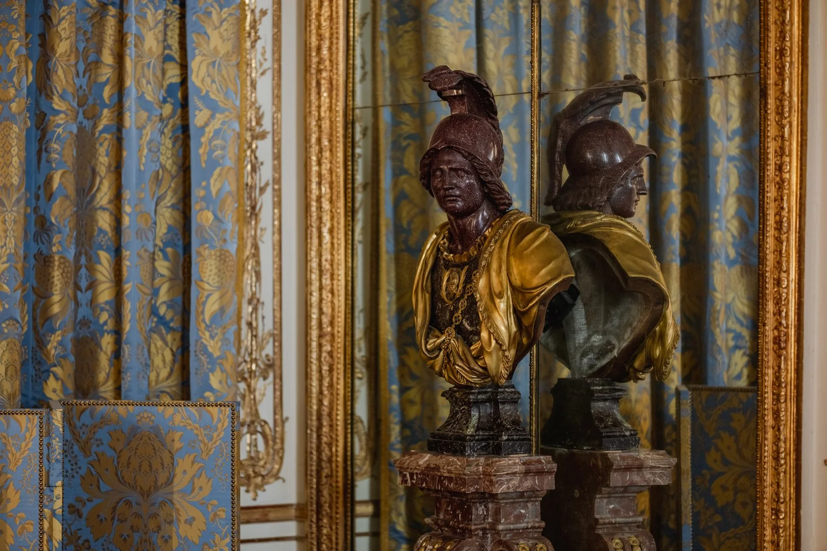 Two classical bust sculptures of Roman soldiers in armor and helmets, displayed on marble pedestals against a background of ornate gold framed mirrors and richly decorated blue and gold patterned curtains.