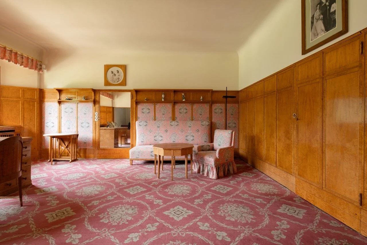 A vintage-style living room with wood-paneled walls and pink patterned carpet. There is a pink upholstered armchair, small wooden tables, and a framed black-and-white photo hanging on the wall.