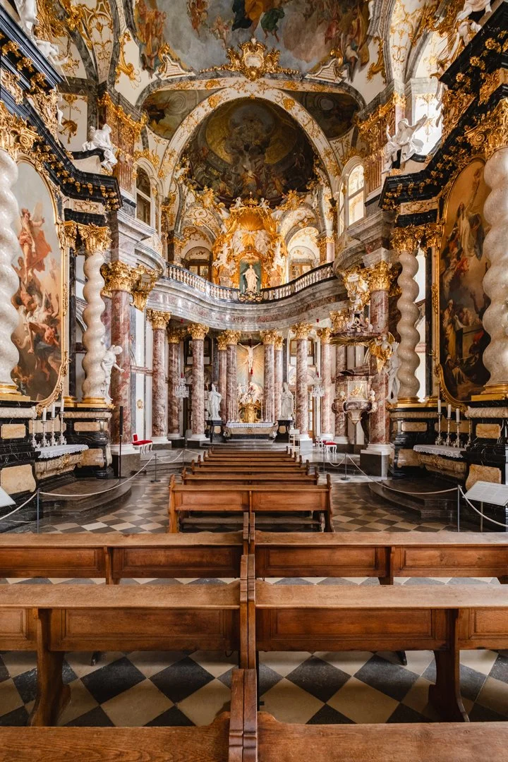 Interior of a Baroque-style church or cathedral with intricate gold and marble decorations, large paintings, and wooden pews facing an altar.