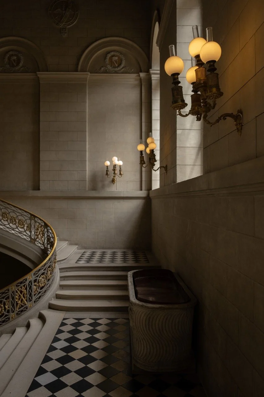 Elegant staircase and balcony inside a historic building with ornate gold railings, wall-mounted lamps with frosted glass globes, surrounded by stone walls and high arched windows.