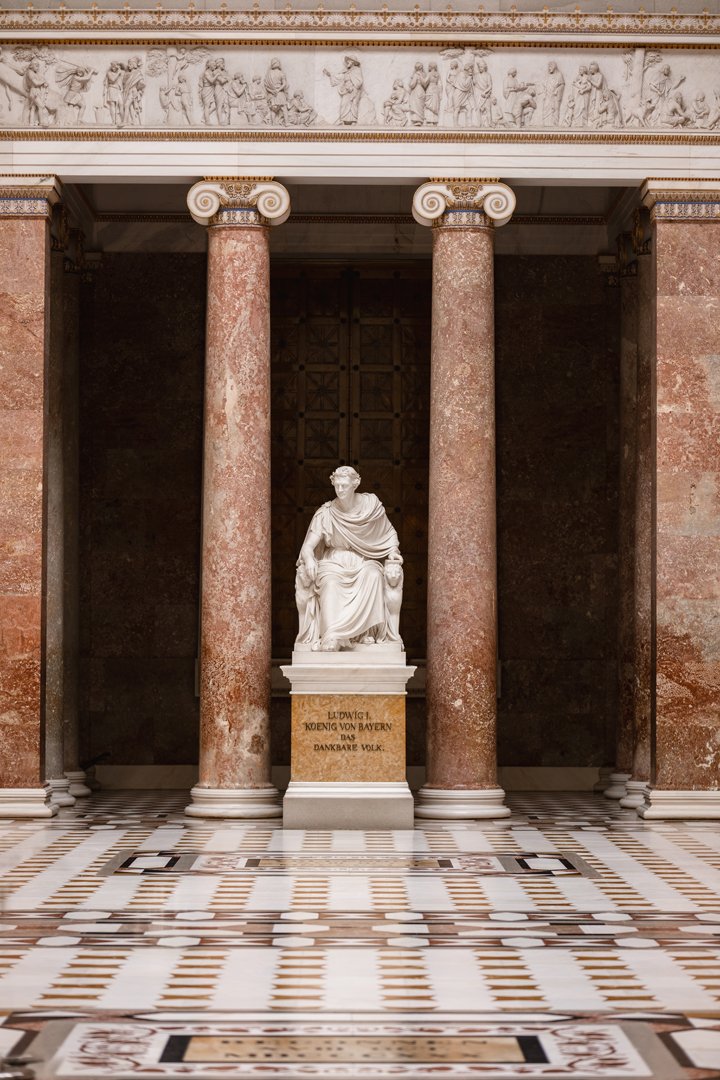 A classical marble statue of a seated man, placed on a pedestal with an inscription, flanked by massive pink marble columns, inside a grand building with ornate architectural details.