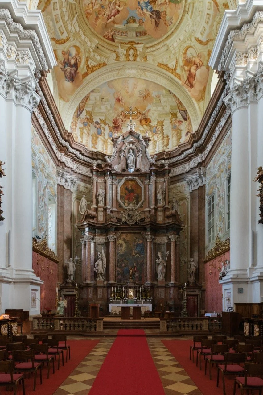 Interior of a Baroque-style church with ornate altar featuring paintings, statues, and columns, red carpet aisle, and ceiling frescoes.
