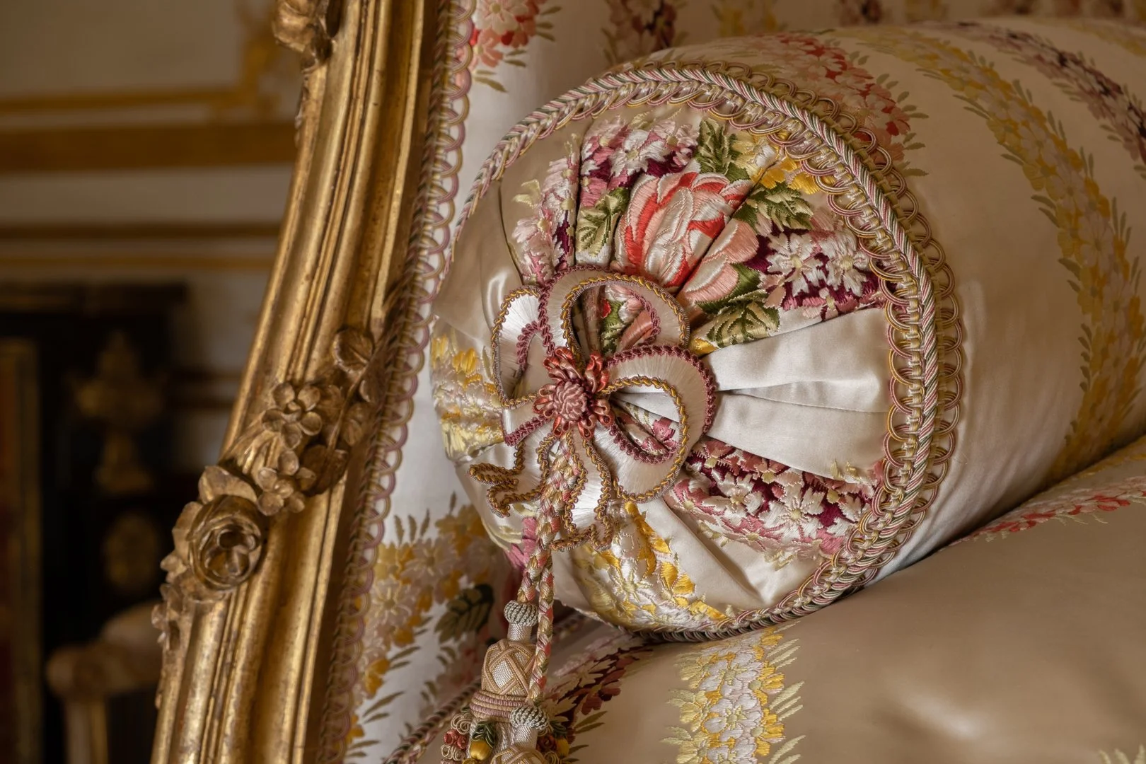 An ornate, embroidered pillow with floral patterns and a decorative tassel and bow, placed on a matching bed with floral upholstery and a gilded frame.