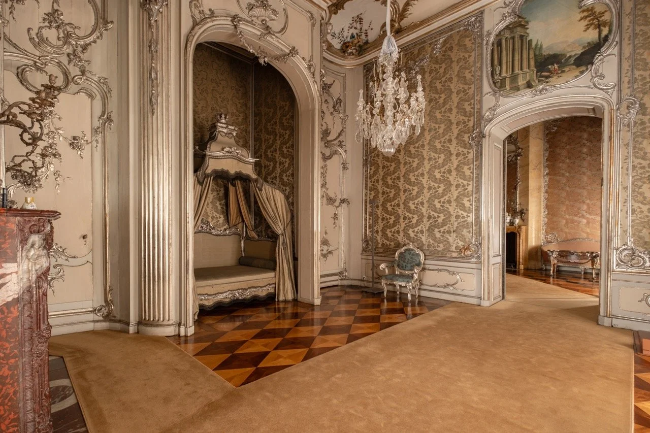 Opulent 18th-century style room with ornate walls, chandelier, small gold and cream daybed with canopy, antique chair, and patterned hardwood and carpet floors.