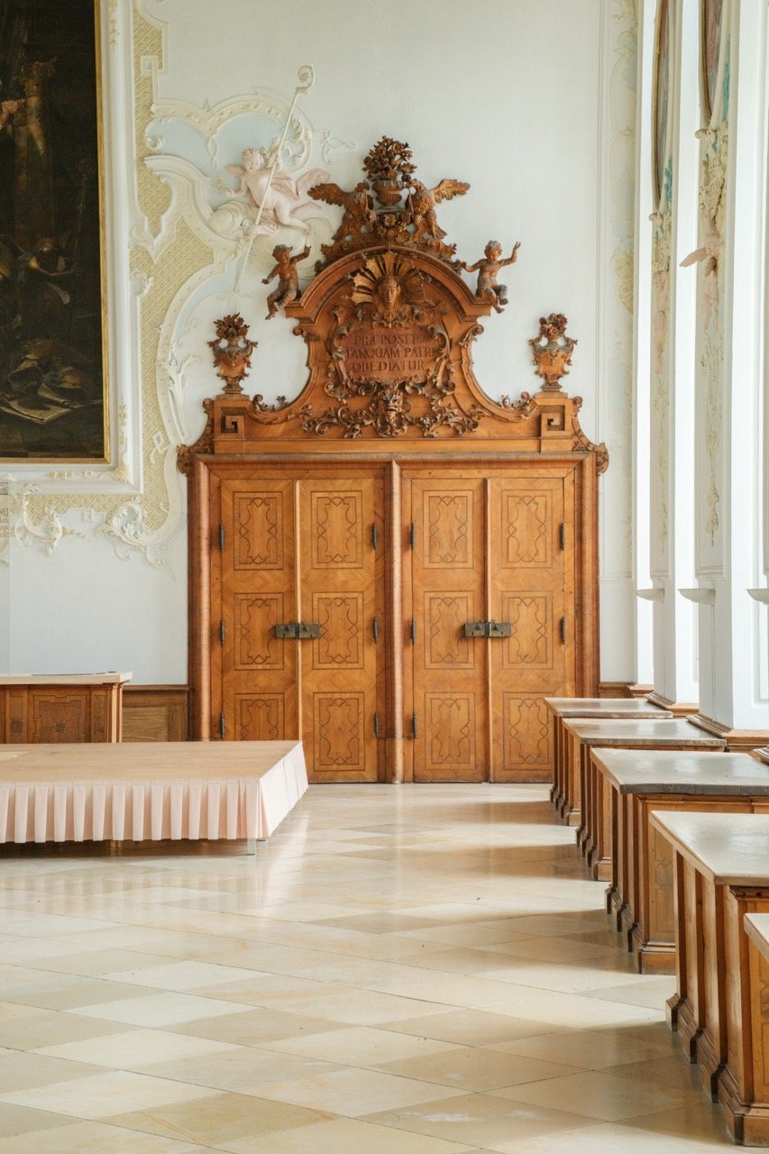 Elegant wooden double doors with ornate carvings and brass hinges, set in a cream-colored wall decorated with intricate moldings and sculptures, in a historic interior room.