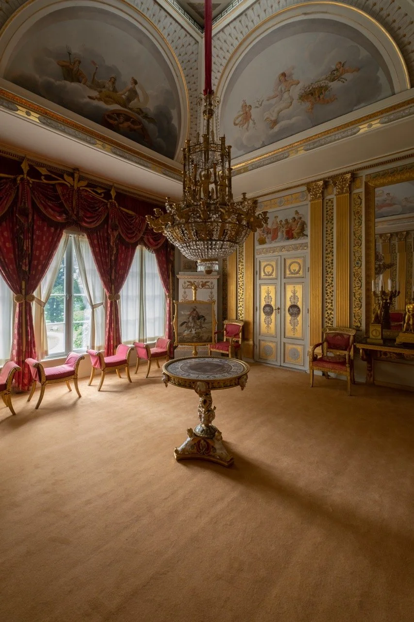 An elegant, ornately decorated room with large windows, red drapes, and pink velvet chairs. The ceiling features elaborate frescoes and a grand chandelier. The room has gold accents, columns, and a small central table with detailed carvings.