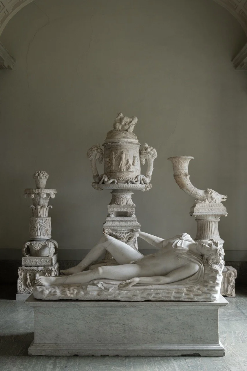 Marble sculpture of a reclining woman lying on her side, with decorative pillars and urns in the background.