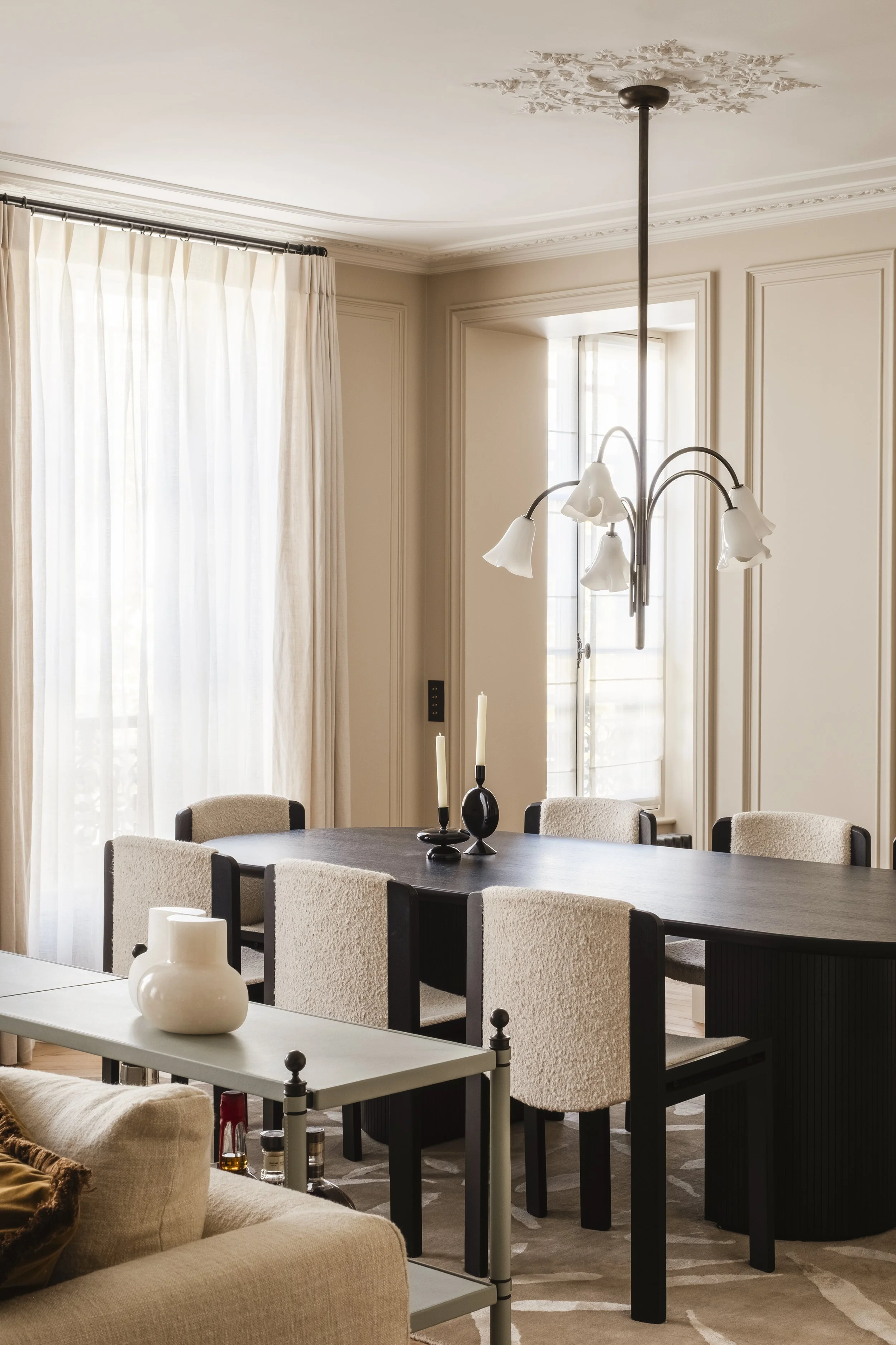 A dining room with a black rectangular table surrounded by six cream and black upholstered chairs. Light-colored curtains cover a window, and a chandelier with five white lampshades hangs above the table. A white side table with a curved black vase a
