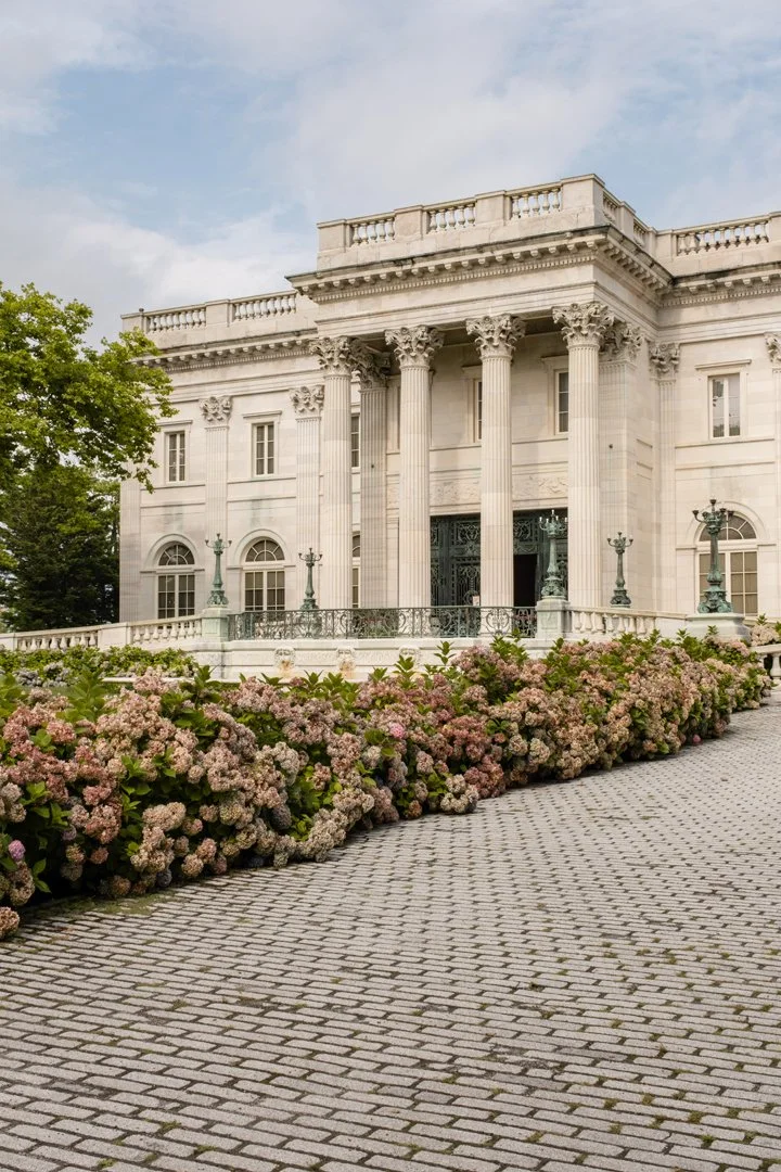 Front view of a grand neoclassical white building with tall columns, decorative balustrades, and lanterns, surrounded by lush greenery and pink flowering bushes, under a partly cloudy sky.