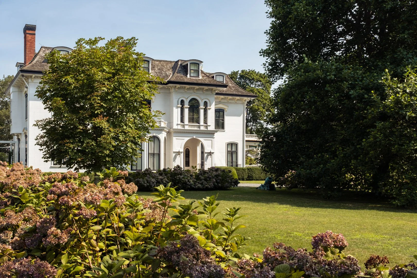 A large white mansion with a steep roof, multiple windows, and an arched entrance, surrounded by green trees, bushes, and a well-maintained lawn on a sunny day.
