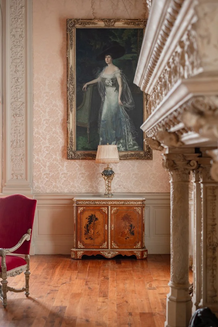 A room with pink damask wallpaper, a large framed portrait of a woman in a black hat hung on the wall, an ornate wooden cabinet with floral and landscape designs, a table lamp on top, a marble fireplace with detailed carvings, a red upholstered armch