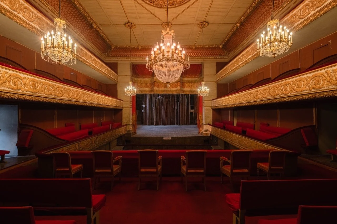 An ornate theater with red velvet seats, gold embellishments, and chandeliers, viewed from the stage.