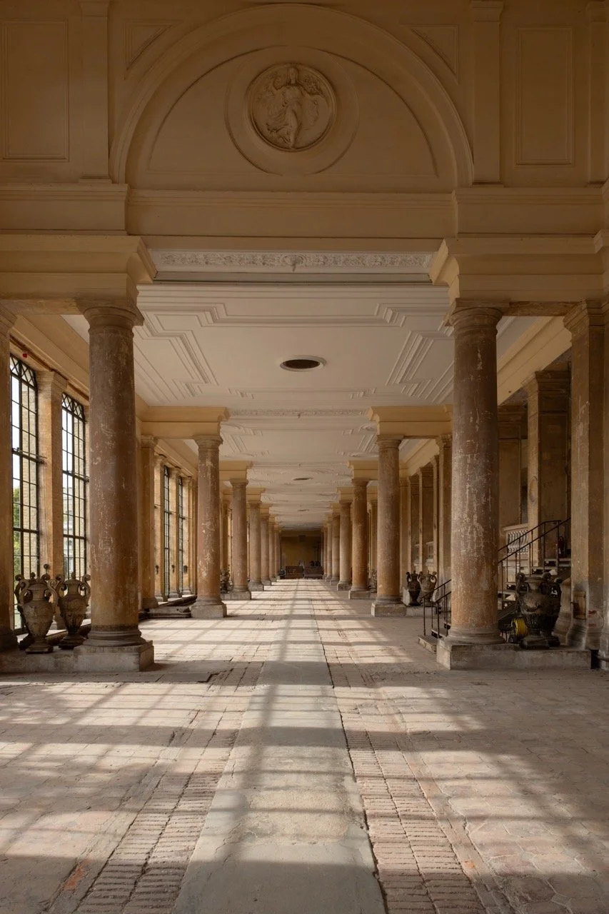 A grand, empty corridor with tall columns, large windows, and decorative ceiling moldings, illuminated by sunlight.