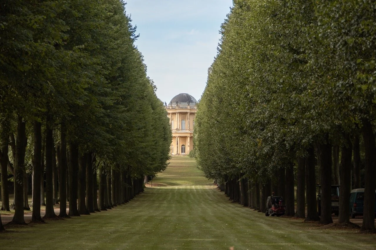 Tree-lined lawn leading to a grand historic building with a domed roof in the distance.