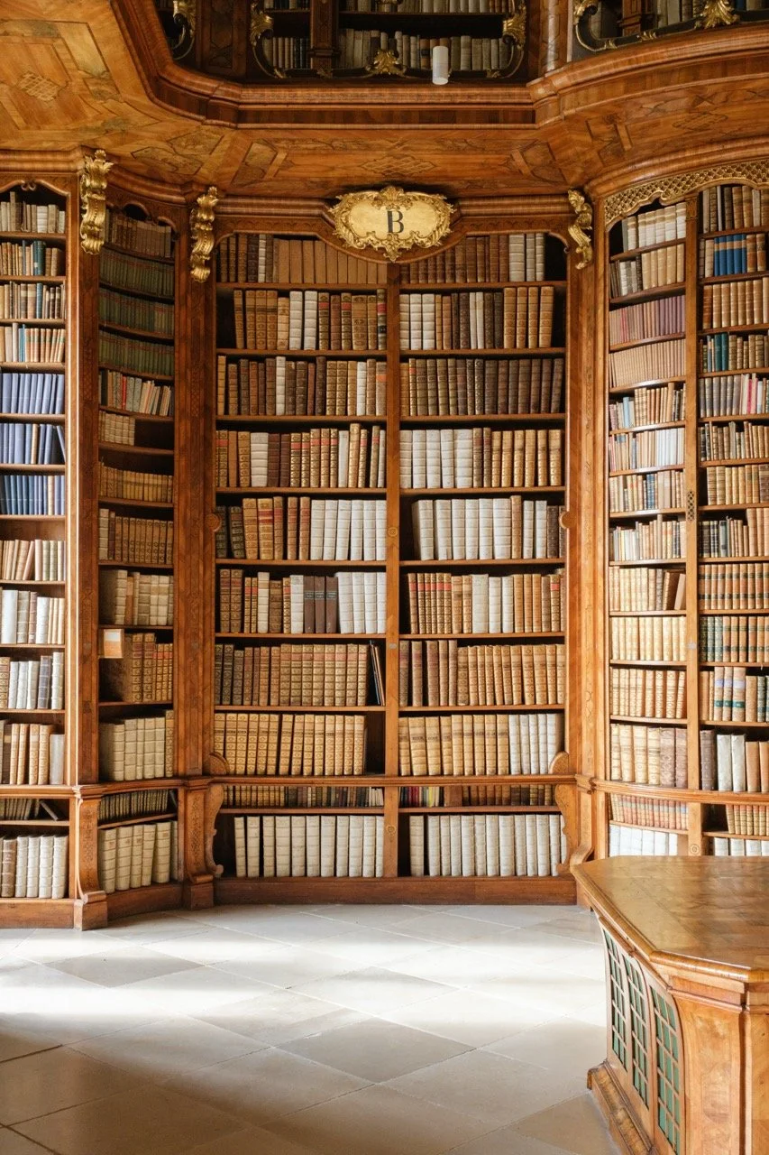 A large, ornate wooden library with tall shelves filled with old books and a wooden table in the foreground.