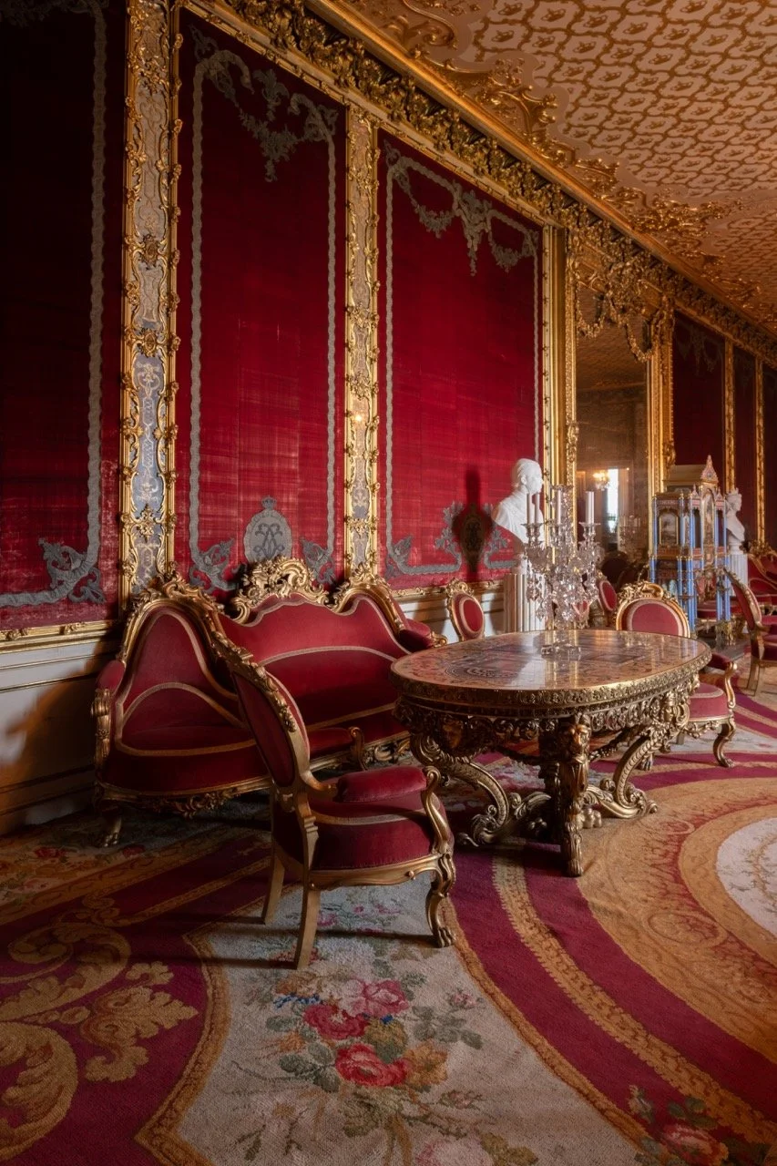 Luxurious interior with red velvet drapes, ornate gold frames, and antique-style furniture including chairs and a table, in a richly decorated room with a patterned carpet.