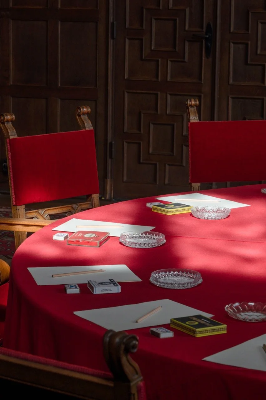 A round table draped with a red tablecloth, set with four white placemats, each with a wooden chopstick and an ashtray, surrounded by red velvet chairs, in front of wooden paneled walls.