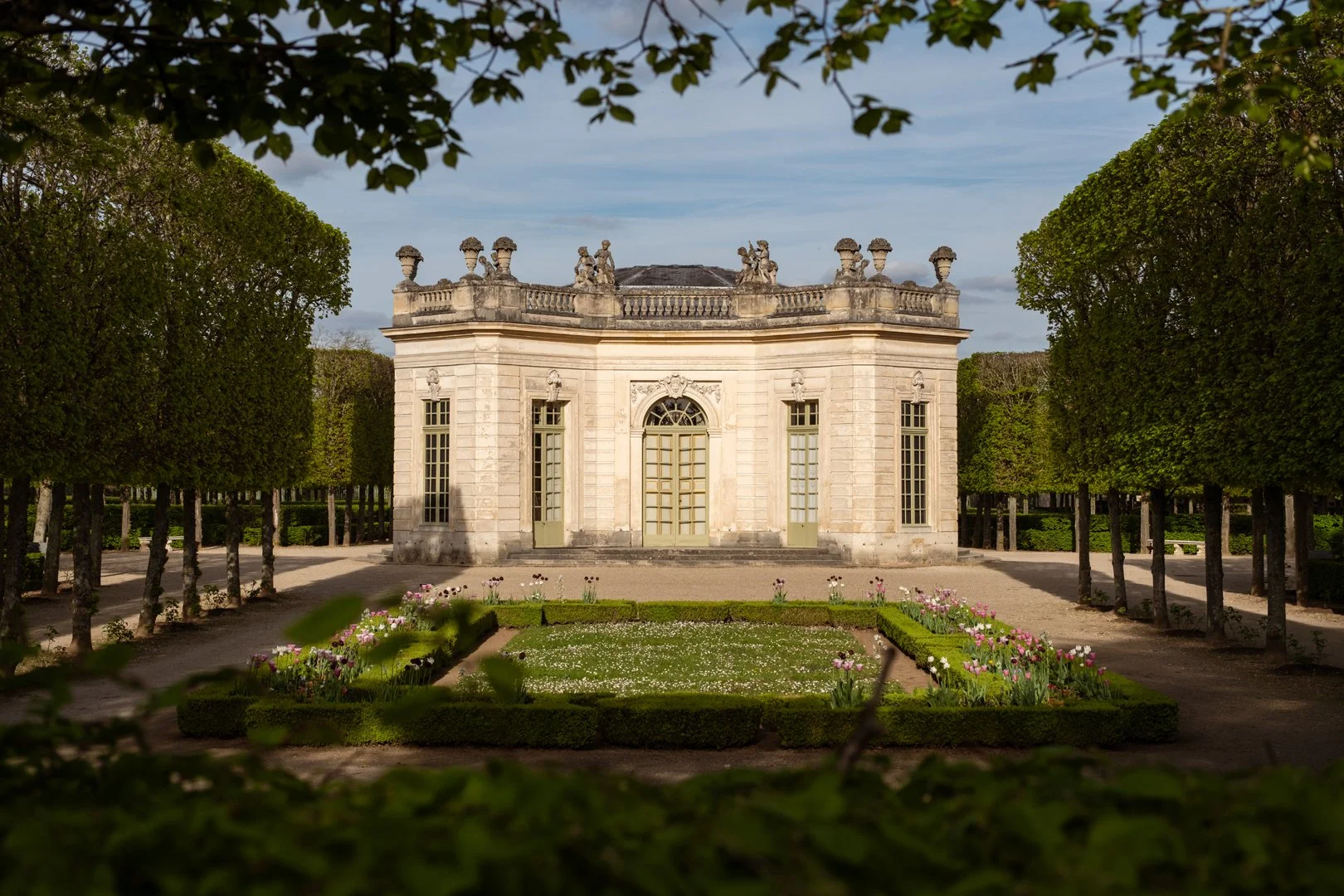 A historic stone building with ornate architectural details, surrounded by manicured gardens and pathways, framed by lush green trees.