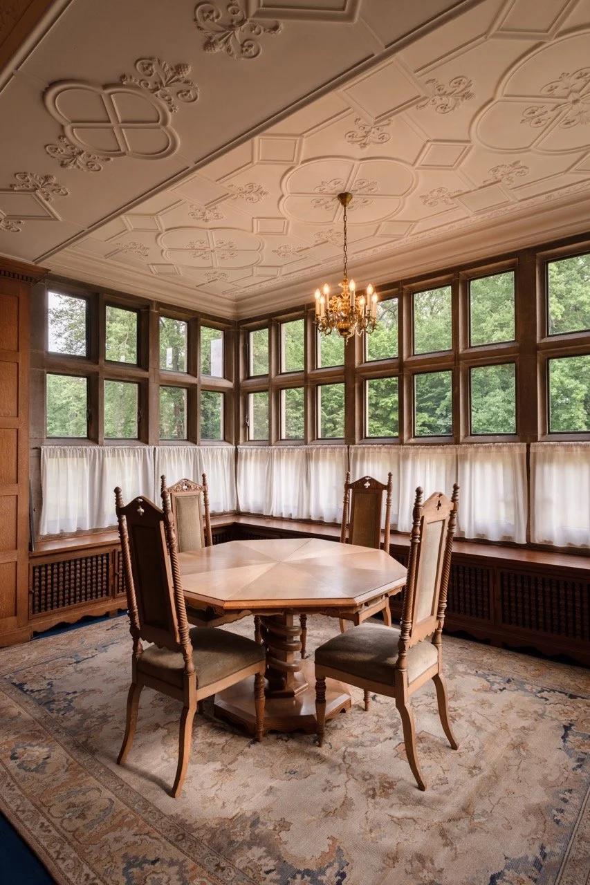 A vintage dining room with a wooden table and four matching chairs, surrounded by large bay windows with white curtains, a chandelier hanging from an ornate ceiling, and a patterned area rug.