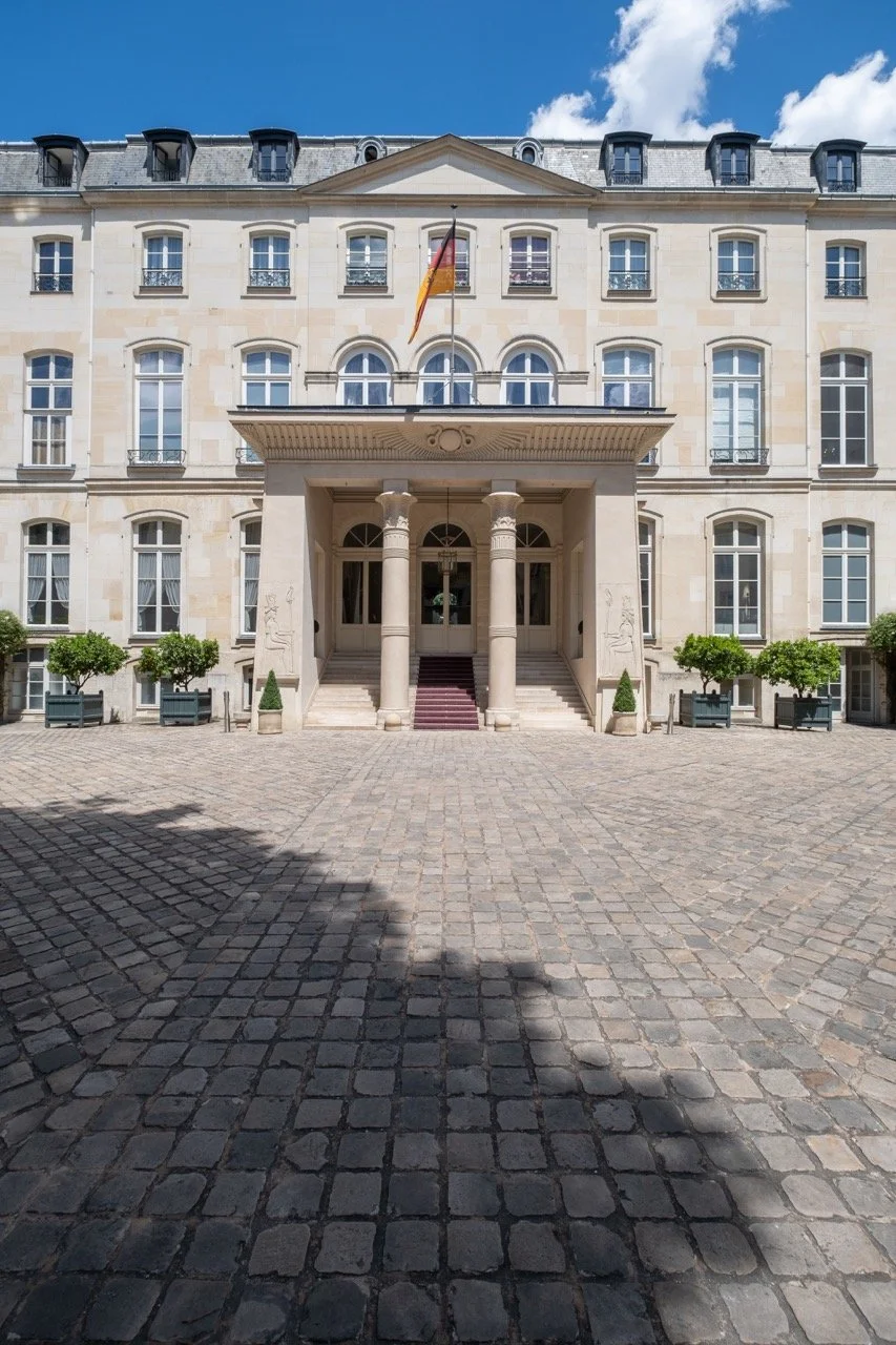 Front view of a historic building with a grand entrance supported by four columns, a cobblestone courtyard, and potted trees along the sides. The building has multiple windows, a flagpole with a flag, and a blue sky with clouds above.