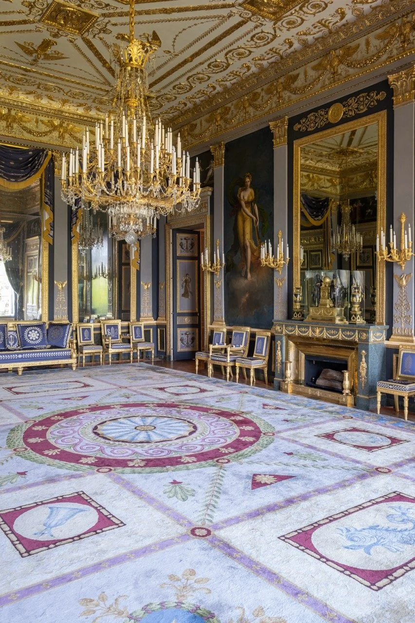 An ornate, luxurious room with a large crystal chandelier, gold-framed mirrors, and classical artwork, featuring a detailed patterned carpet and blue velvet curtains.