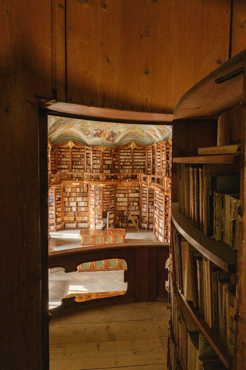 A view of a grand, circular library with wooden bookshelves filled with books, taken from a small, curved balcony with a wooden railing, and decorated with ornate, golden accents and a painted ceiling.