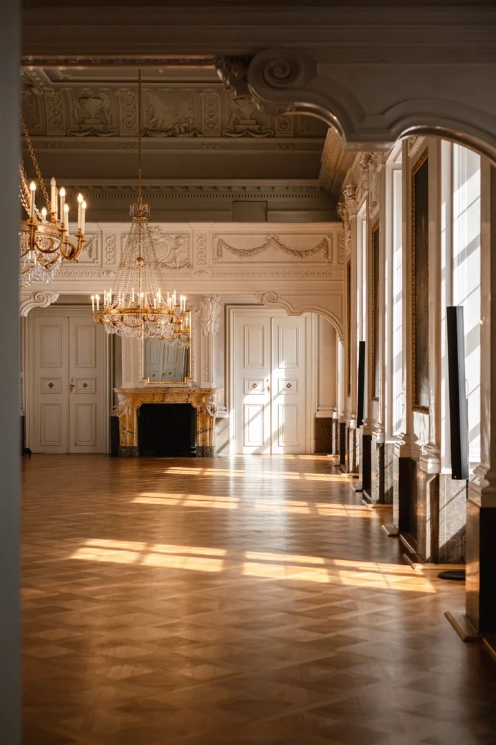 Elegant, empty grand ballroom with ornate white walls, decorative moldings, chandeliers, a fireplace, large windows with curtains, and wooden parquet flooring illuminated by sunlight.
