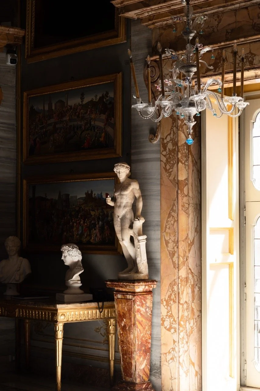 Interior of a luxurious room featuring classical sculptures, marble columns, a chandelier, and framed paintings.