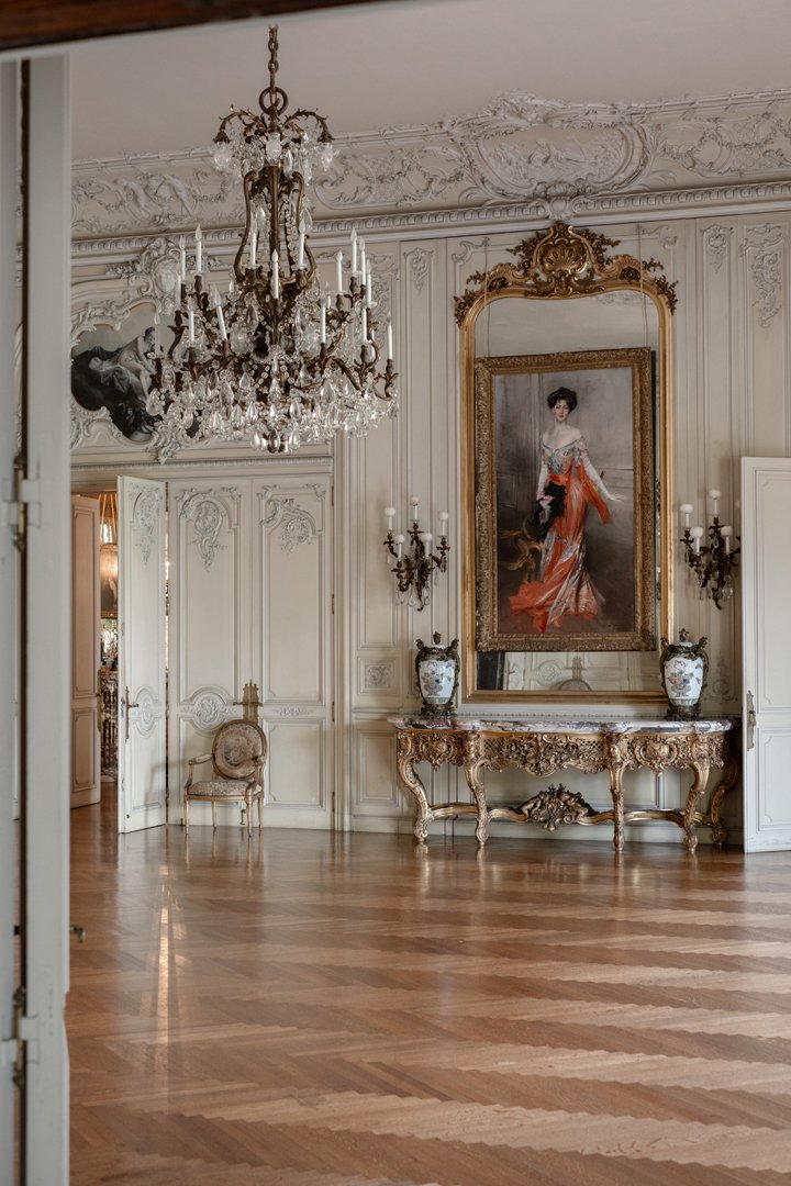 Elegant room with a grand crystal chandelier, ornate white walls, a large framed portrait of a woman in a flowing dress, and a decorative console table with vases.