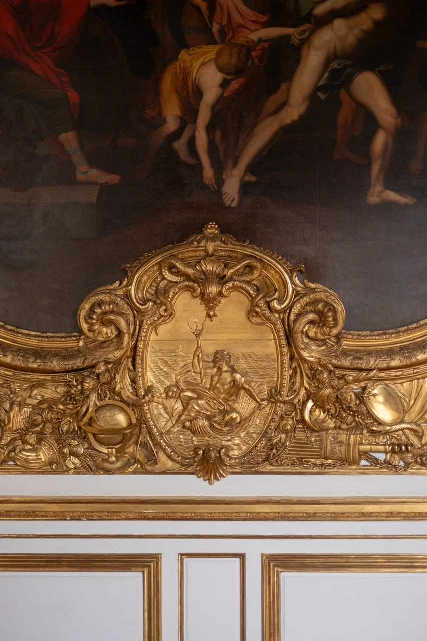 Close-up of a baroque-style gold ornamental frame with a classical painting depicting mythological figures, set above a white panel with gold trim.