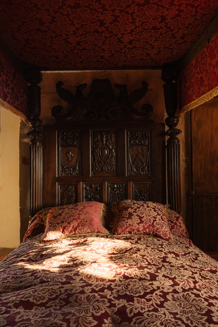 A vintage bed with a dark, ornate wooden headboard and matching pillows, illuminated by sunlight, in a room with red and gold patterned wallpaper and wooden accents.