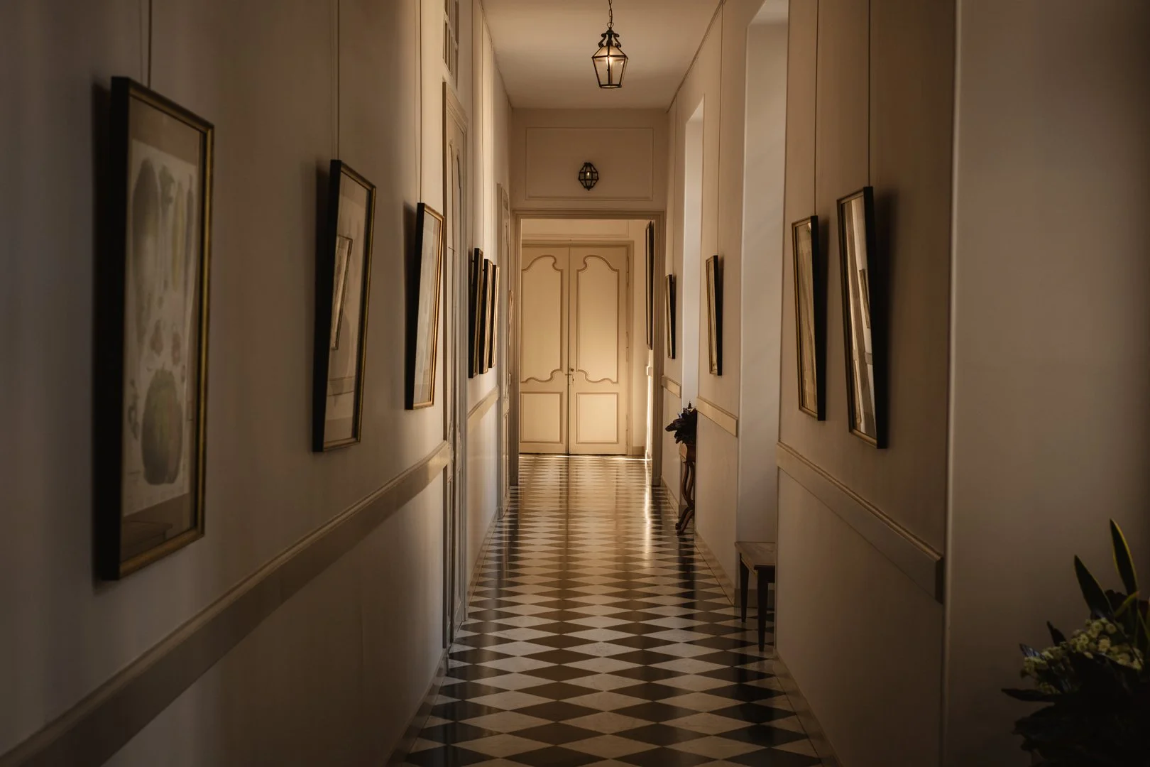 A narrow hallway with black and white checkered floor tiles, lined with framed pictures on both sides, leading to a closed double door at the end, with a hanging ceiling light and a small table with a flower arrangement.