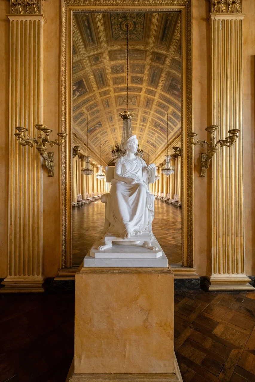 A white marble sculpture of a seated woman holding a book, placed on a pedestal in a grand, ornately decorated hall with gold accents, chandeliers, and a reflection of the hall's ceiling in a large mirror behind the statue.