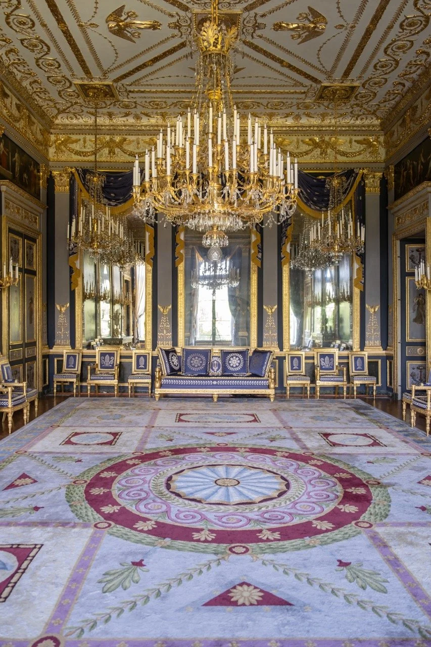An opulent room with elaborate gold accents, a large chandelier, gilded chairs with blue upholstery, mirrored walls, and a decorative carpet with floral and geometric patterns.