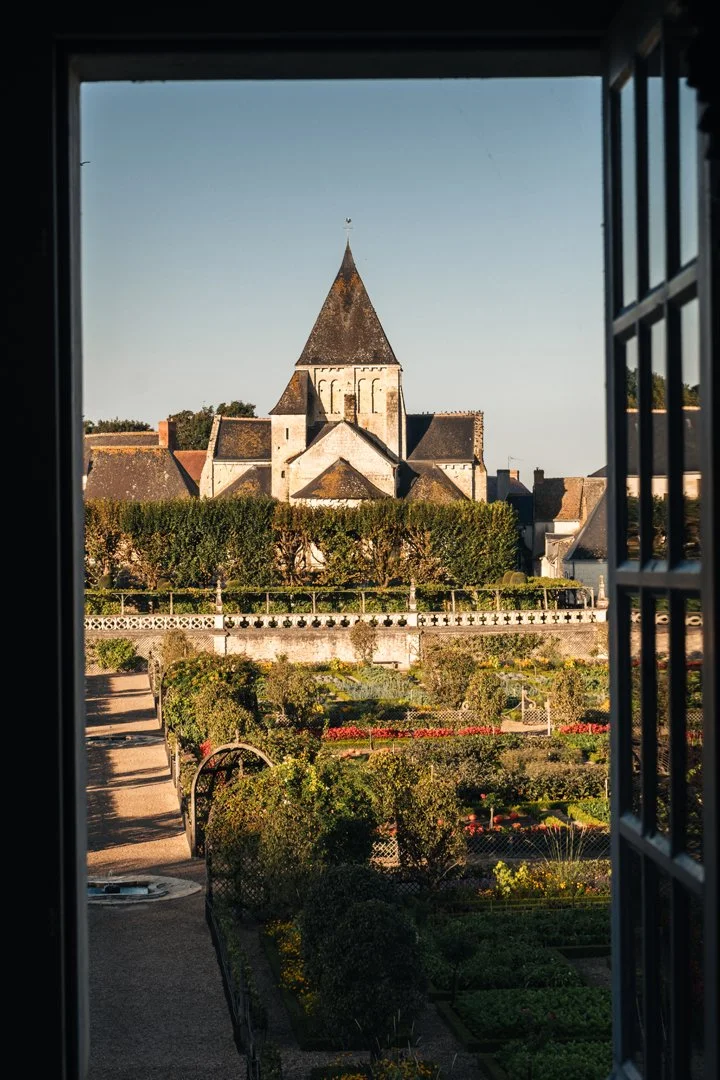 View of a historic stone church with a pointed steeple seen through an open window, overlooking a well-maintained garden with hedges and flowerbeds.