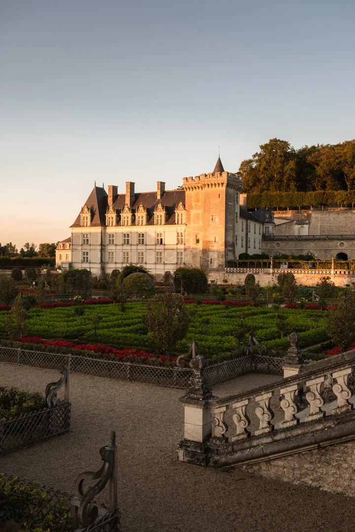 A historic castle with multiple towers and steep rooftops illuminated by sunset, surrounded by manicured gardens with colorful flowerbeds and decorative stone railings.