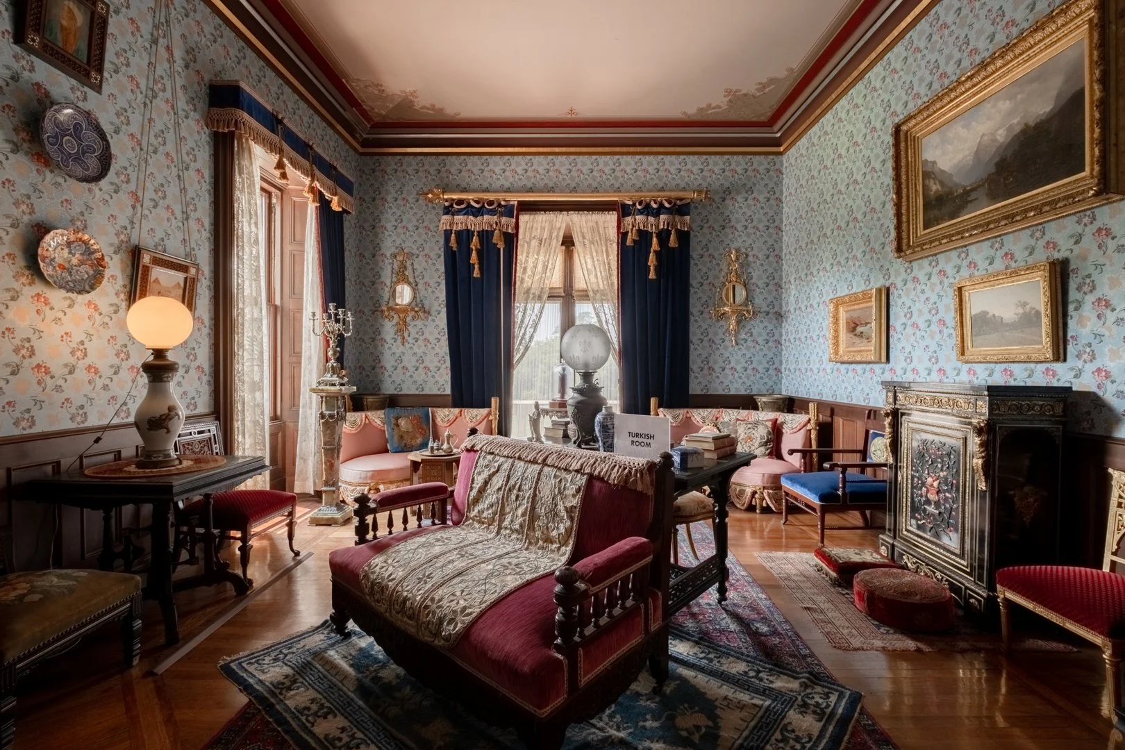 A vintage sitting room decorated with floral wallpaper, wooden flooring, and antique furniture including sofas, armchairs, tables, and a fireplace. There are framed paintings on the walls, ornate curtains on the windows, and decorative plates on one 