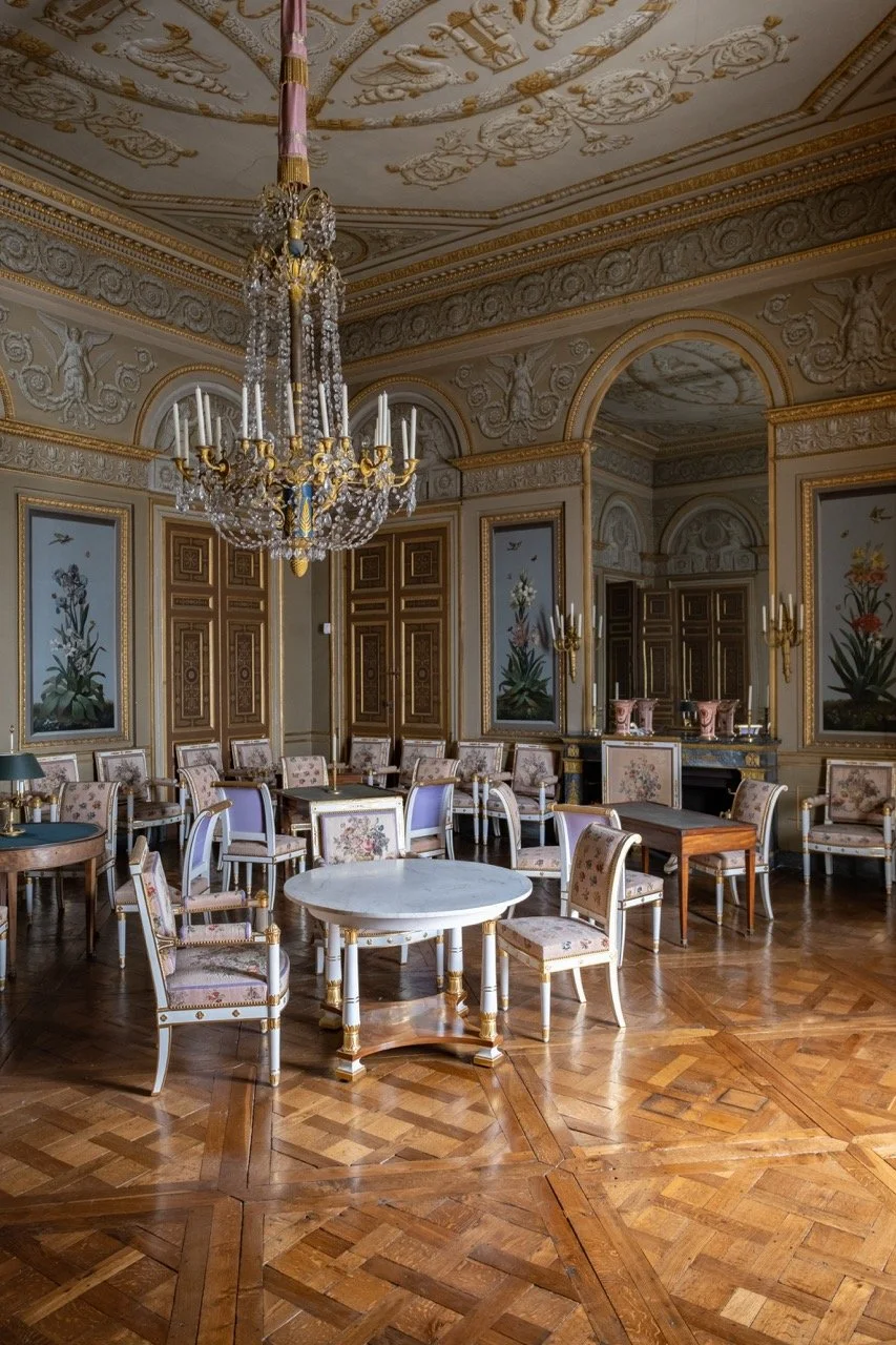 Elegant opulent interior of a room with ornate gold and white decorative moldings, a grand chandelier, floral art on the walls, and vintage-style chairs and tables on a polished wooden floor.