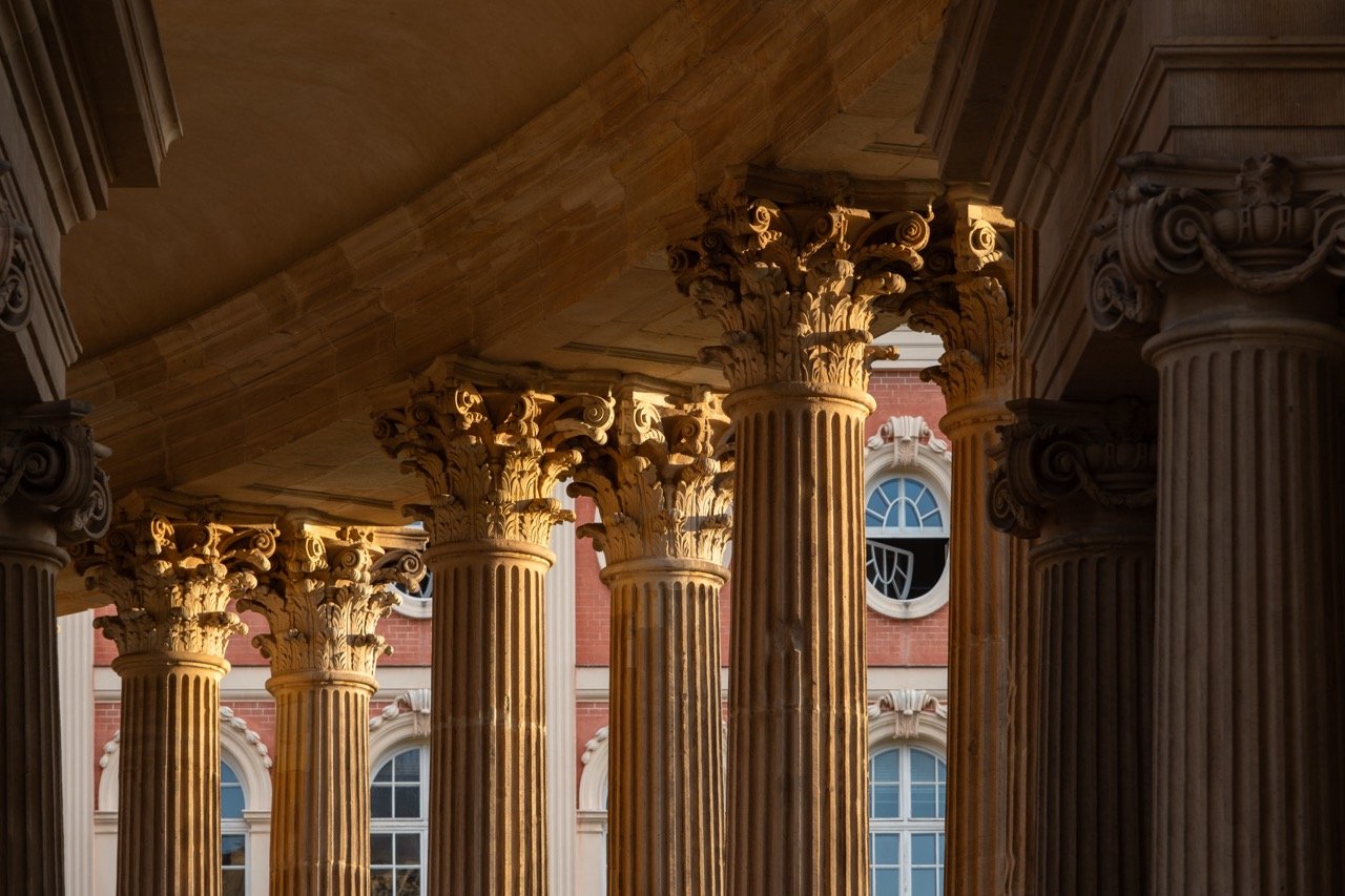 View of classical columns with ornate capitals in front of a red brick building with arched windows.