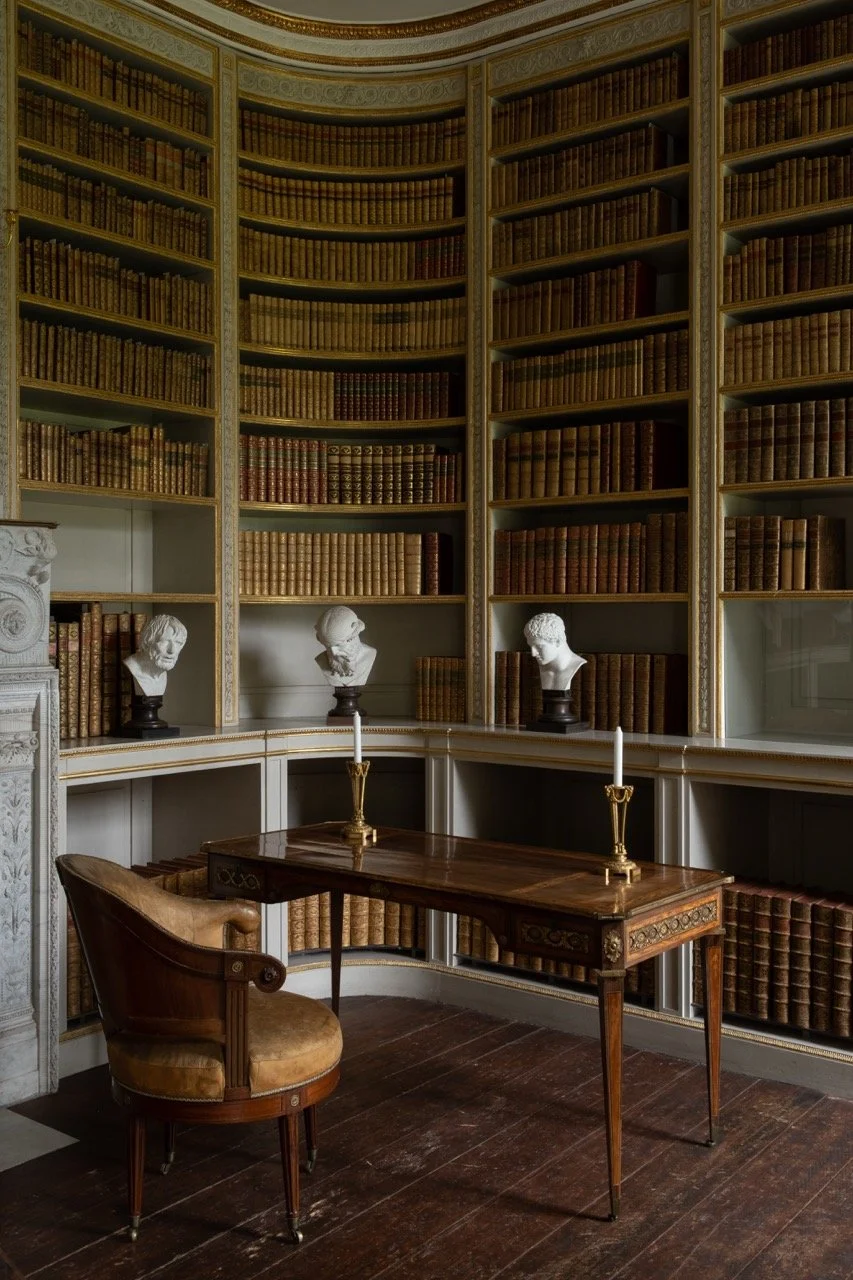 A vintage study room with curved bookshelves filled with old leather-bound books, a wooden desk with two tall candlesticks and a vintage chair, and bust statues on the shelves.