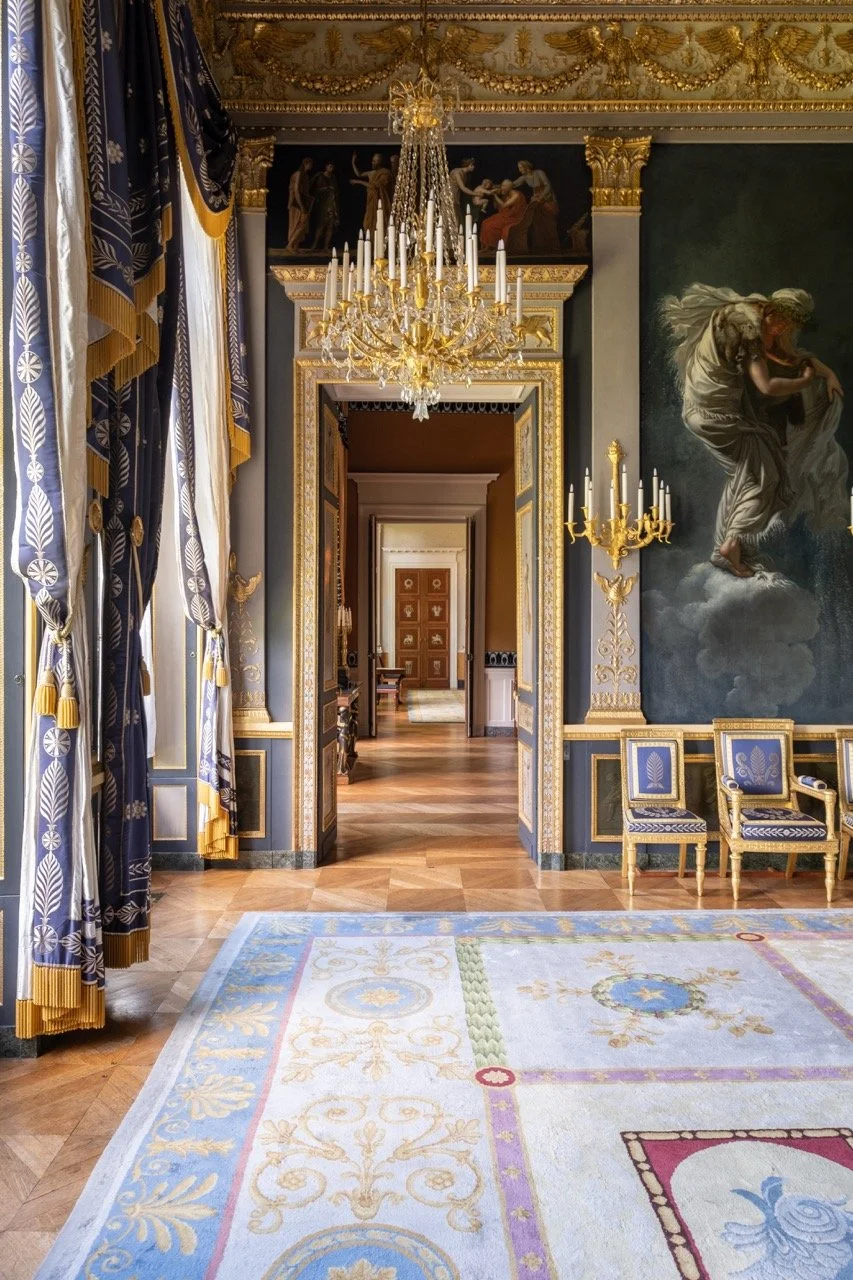 Opulent room with gold accents, large chandelier, ornate curtains, classical wall décor, and a colorful rug.