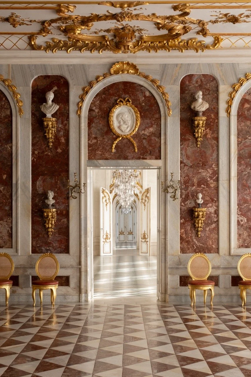 Luxurious room with ornate gold and marble decor, classical bust sculptures in niches, and an open doorway leading to a chandelier-lit hall.