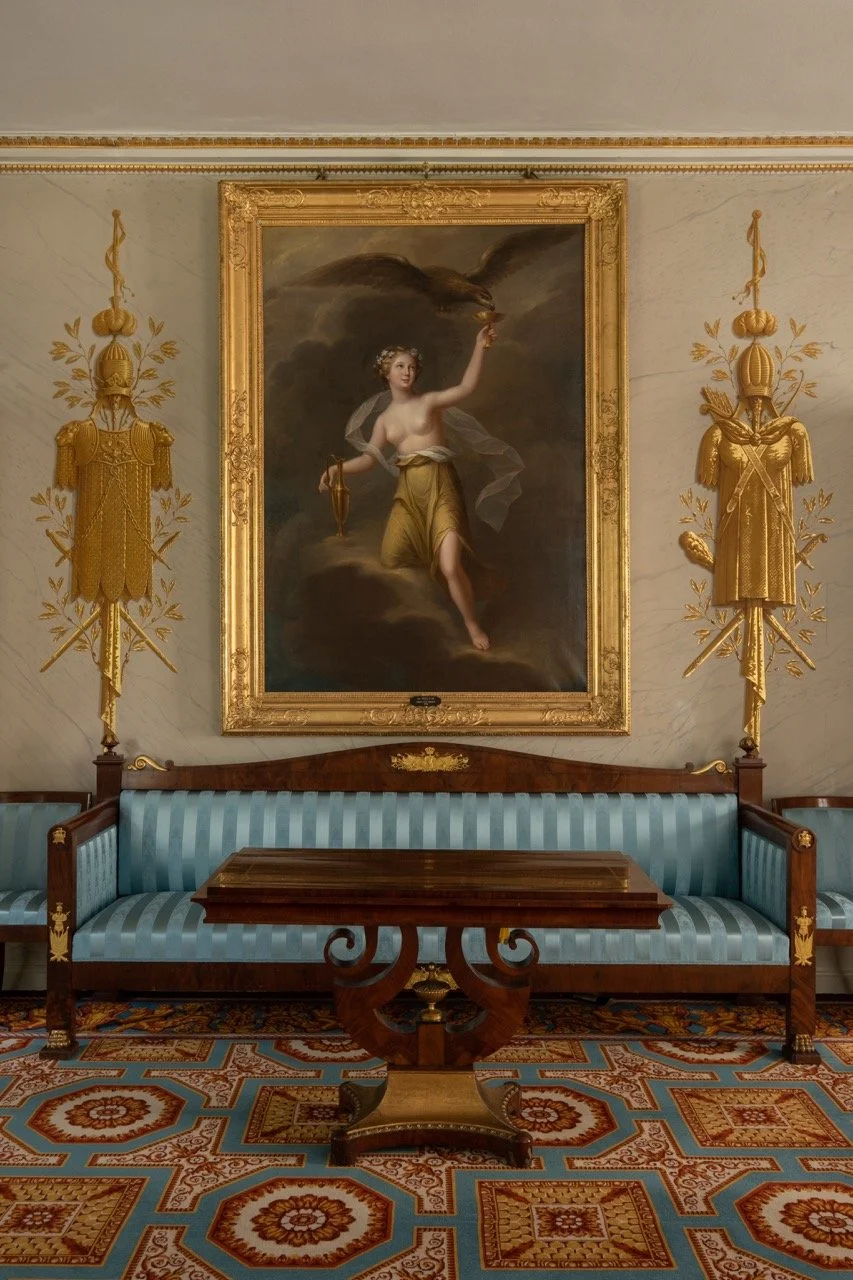 A room with a large framed painting of a woman with a cloud and eagle above her, flanked by gold decorative wall pieces, a blue striped vintage sofa, a wooden coffee table and ornate patterned carpet.