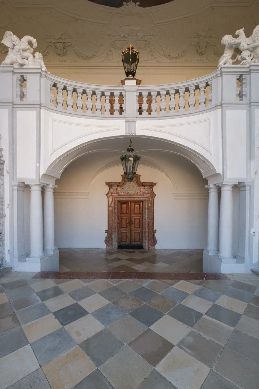 Elegant interior with a wooden door, arching white columns, decorative sculptures, and vintage hanging lanterns.